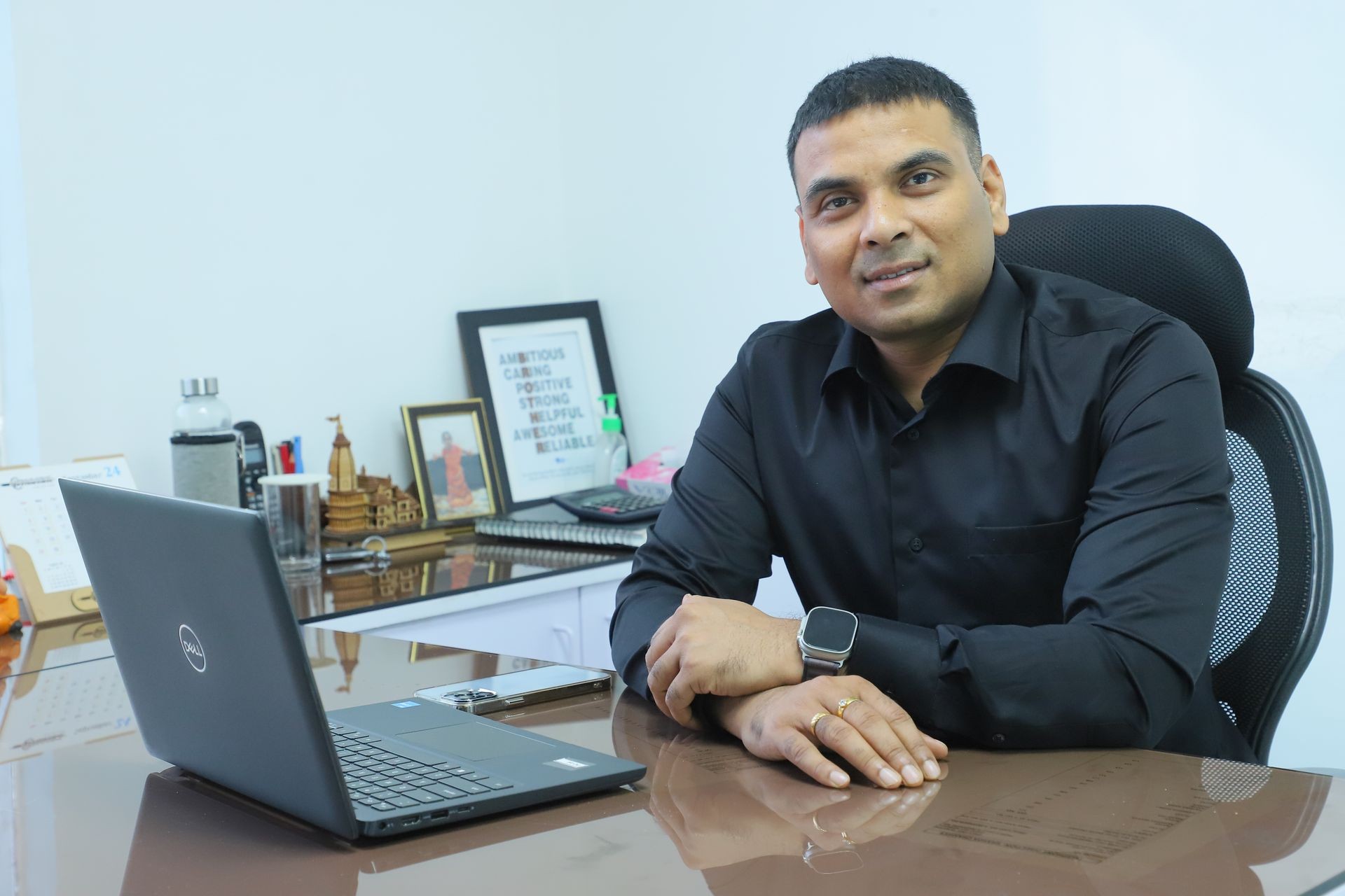 Man in black shirt sitting at a desk with a laptop and framed pictures.