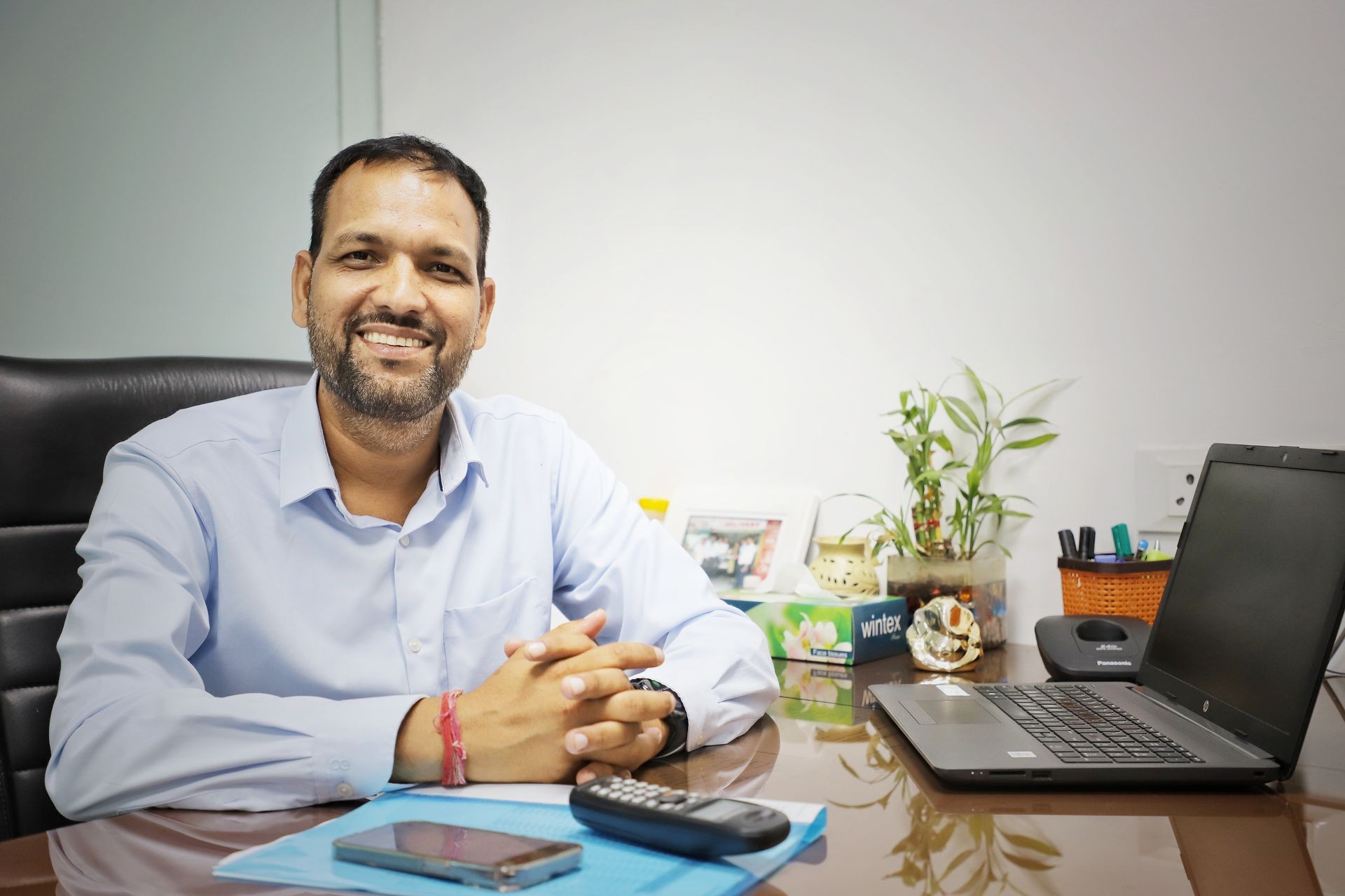 Smiling man in a light blue shirt seated at an office desk with a laptop and plants.