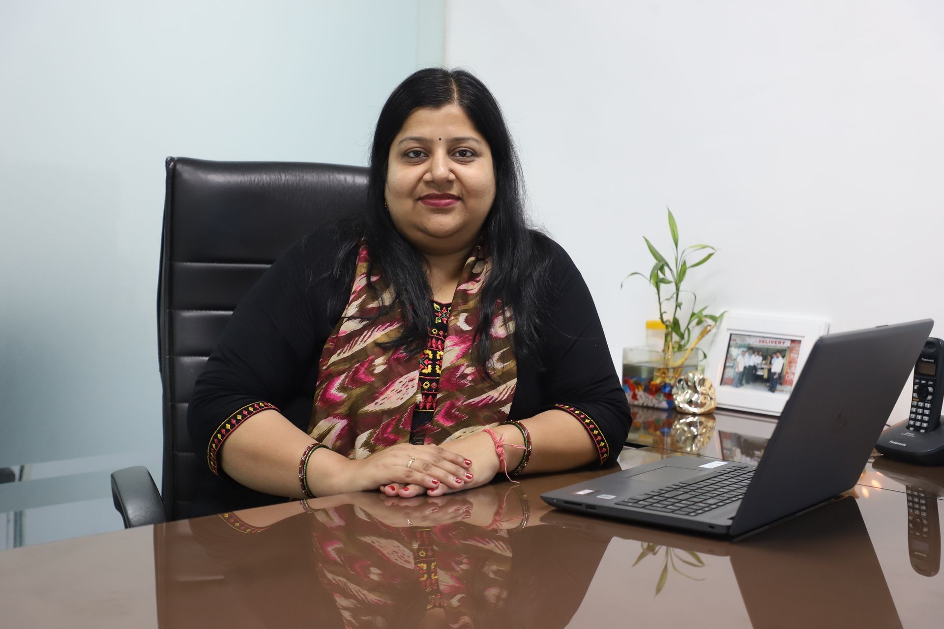 Woman sitting at a desk with a laptop, office plant, and framed photo in the background.